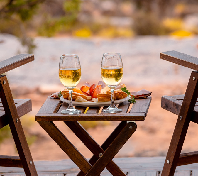 Two glasses of white wine and a plate of fruit on a small wooden table in a desert setting.