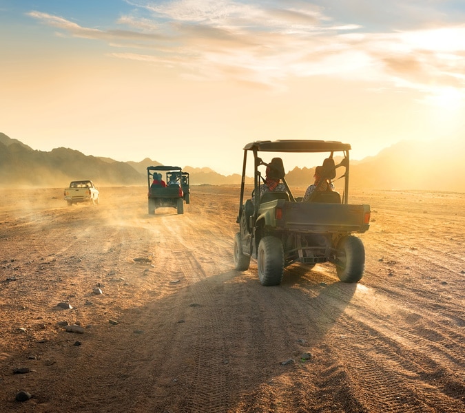 A convoy of off-road vehicles driving across dusty desert terrain with mountains in the background.