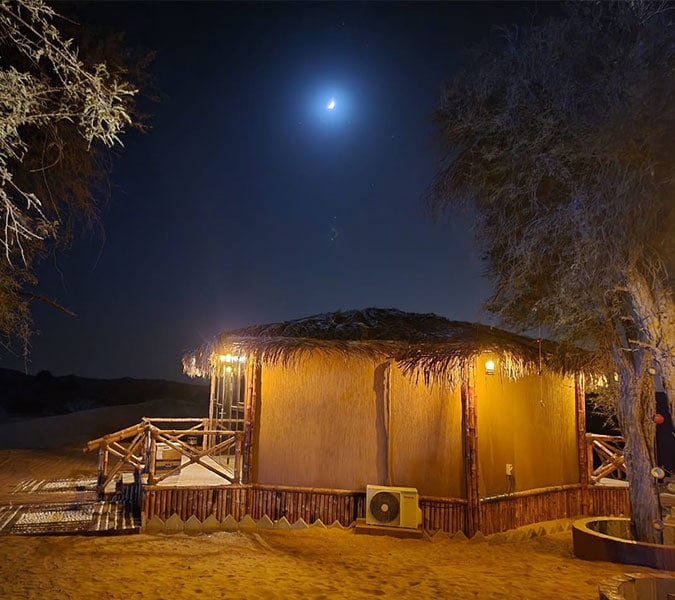 A circular, thatched-roof hut illuminated under a crescent moon in the desert.