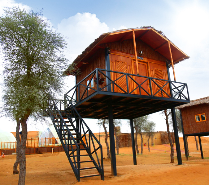 An elevated wooden cabin on stilts with a small balcony, set in a desert camp.
