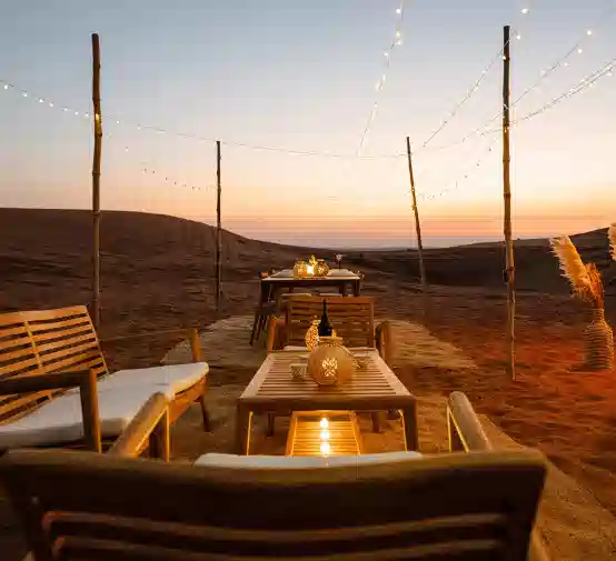 A private desert dining area at dusk, featuring simple wooden tables, soft lights, and a warm sunset backdrop.