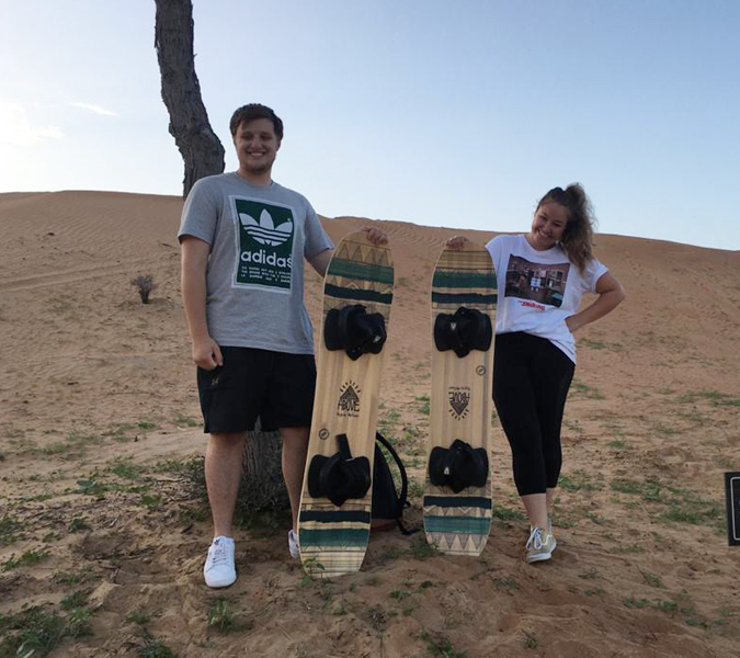 Two people holding sandboards, standing on desert ground with dunes behind them.