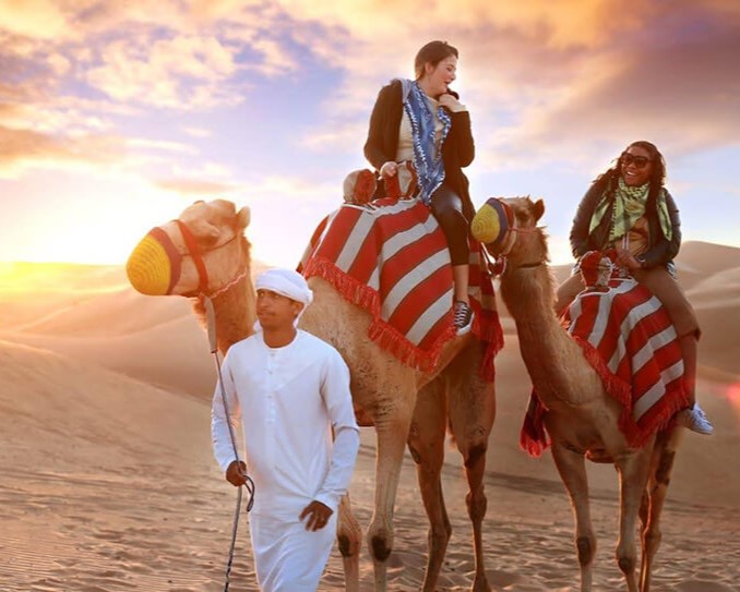 Two travelers on camels guided through the desert at sunset