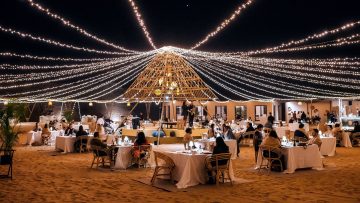 An outdoor desert restaurant setup at night with white-clothed tables on the sand, all under a massive canopy of fairy lights.