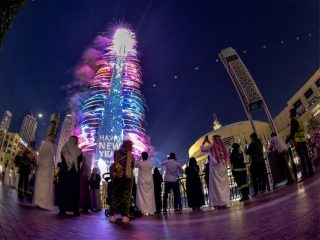 A low-angle fisheye lens photo of a crowd in traditional and modern clothing looking up at a colorful firework display on the Burj Khalifa.