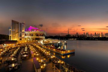 An outdoor restaurant deck with many tables lit by small lamps at dusk, situated on the water with the Dubai skyline and Burj Khalifa on the horizon.
