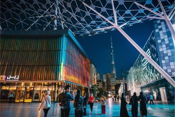 People walking through a modern outdoor shopping area at night under a geometric white canopy, with the Burj Khalifa visible in the distance.