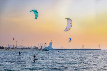 Several kitesurfers and hydrofoilers on the blue ocean with colorful kites in the air and the Burj Al Arab in the distant background under a hazy sunset.